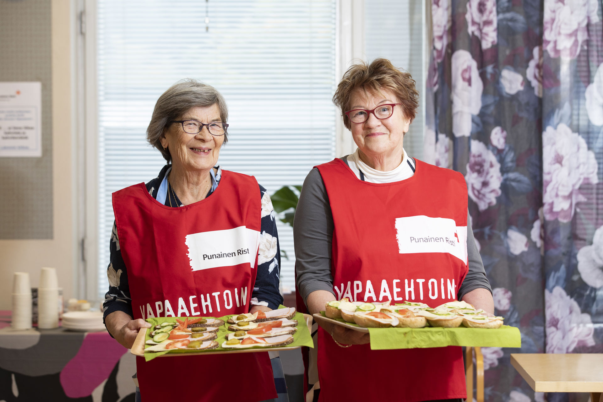 Två frivilliga ler mot kameran och håller brickor med smörgåsar i sina händer.