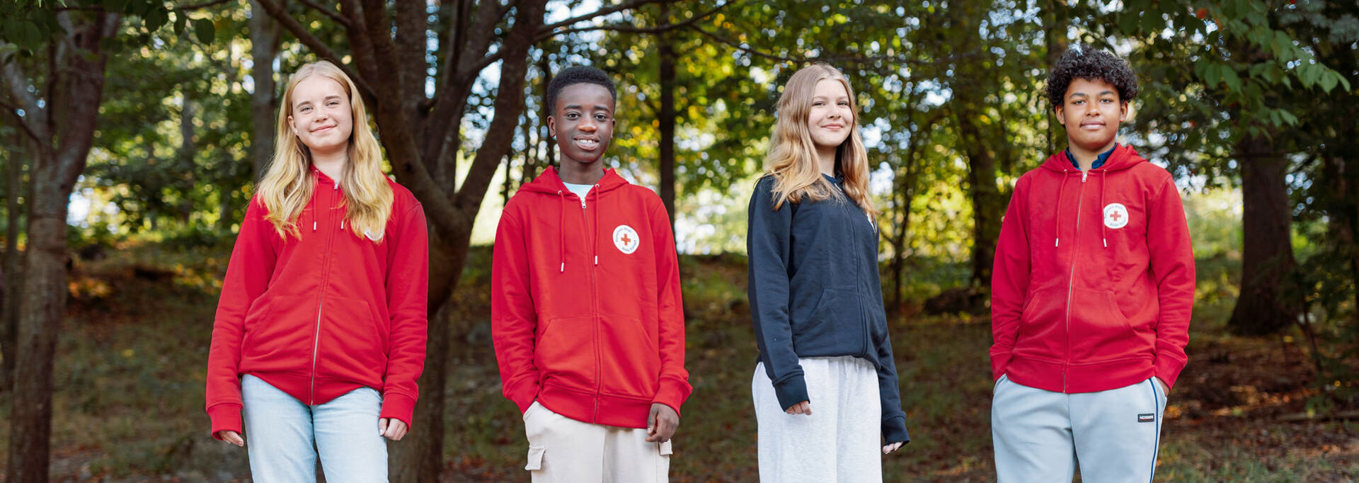 Fyra av Röda Korsets unga frivilliga står utomhus. Tre av dem har på sig Röda Korsets röda huvtröja.