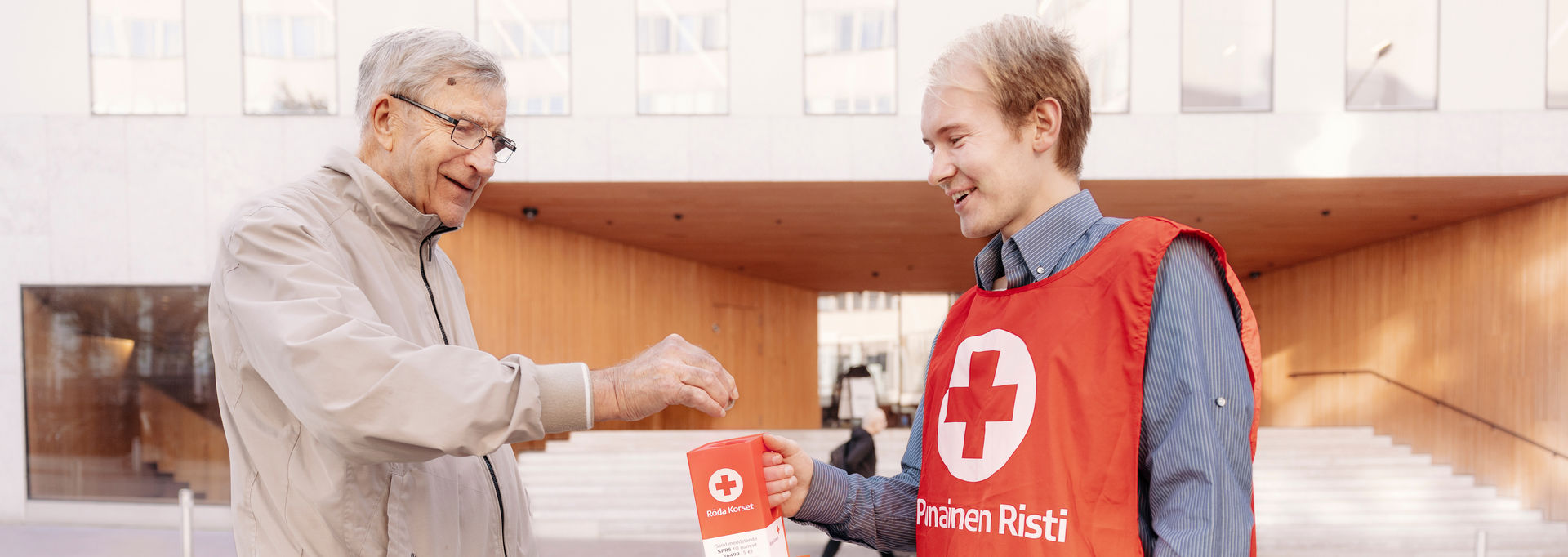 En leende person lägger pengar i en Röda Korsets frivilliga insamlingslåda.