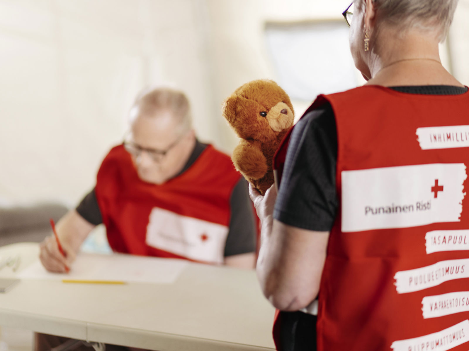 En av nallesjukhusets frivilliga skriver på ett papper i bakgrunden och den andra har en nalle i händerna.