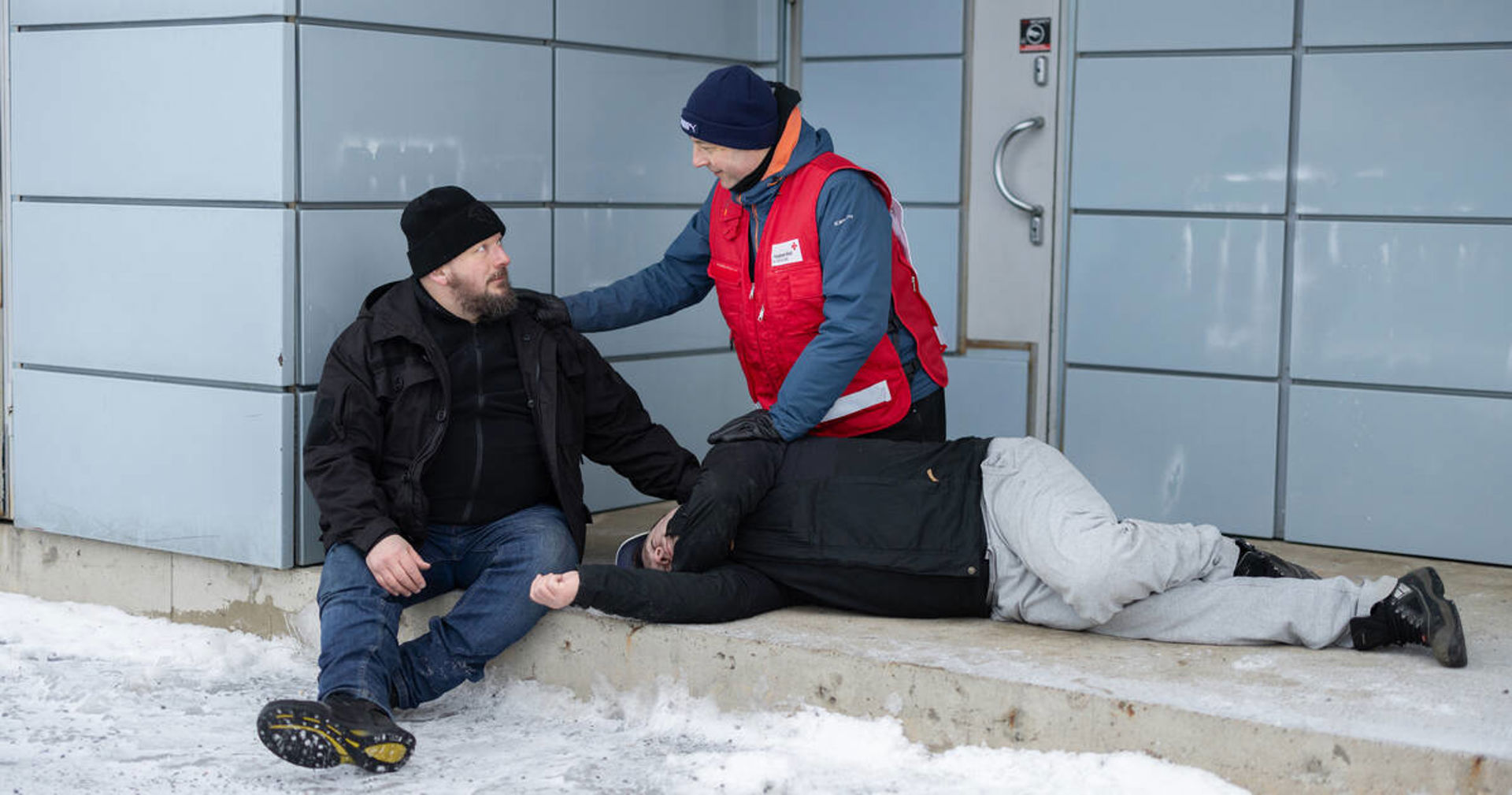 En Röda Kors-frivillig i röd väst hjälper två personer utanför en byggnad under vintriga förhållanden. Den ena personen sitter på ett trappsteg och den andra ligger på sidan på marken.