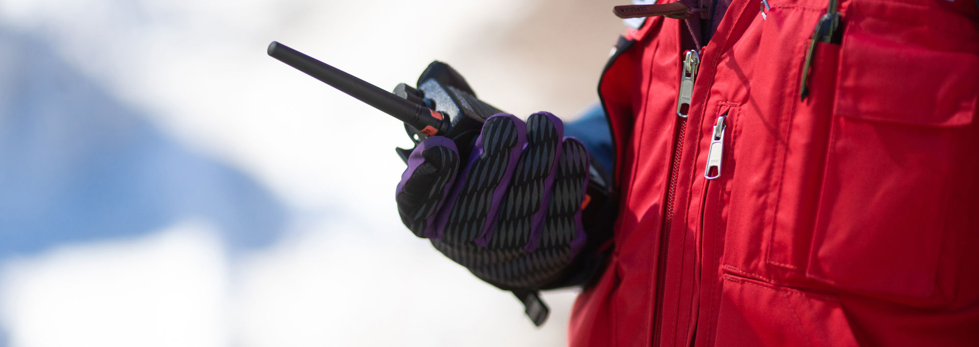 En person klädd i röd väst håller en radiotelefon i sin handskklädda hand. Bakgrunden är vit och snöig.