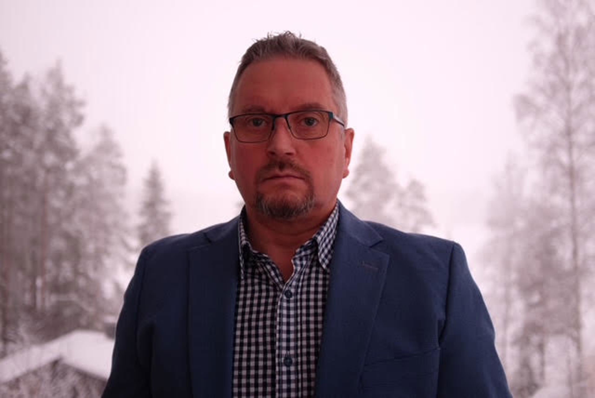 Heikki Väätämöinen tittar mot kameran, klädd i en blå kavaj och rutig skjorta. I bakgrunden finns ett vinterlandskap med snöiga träd.