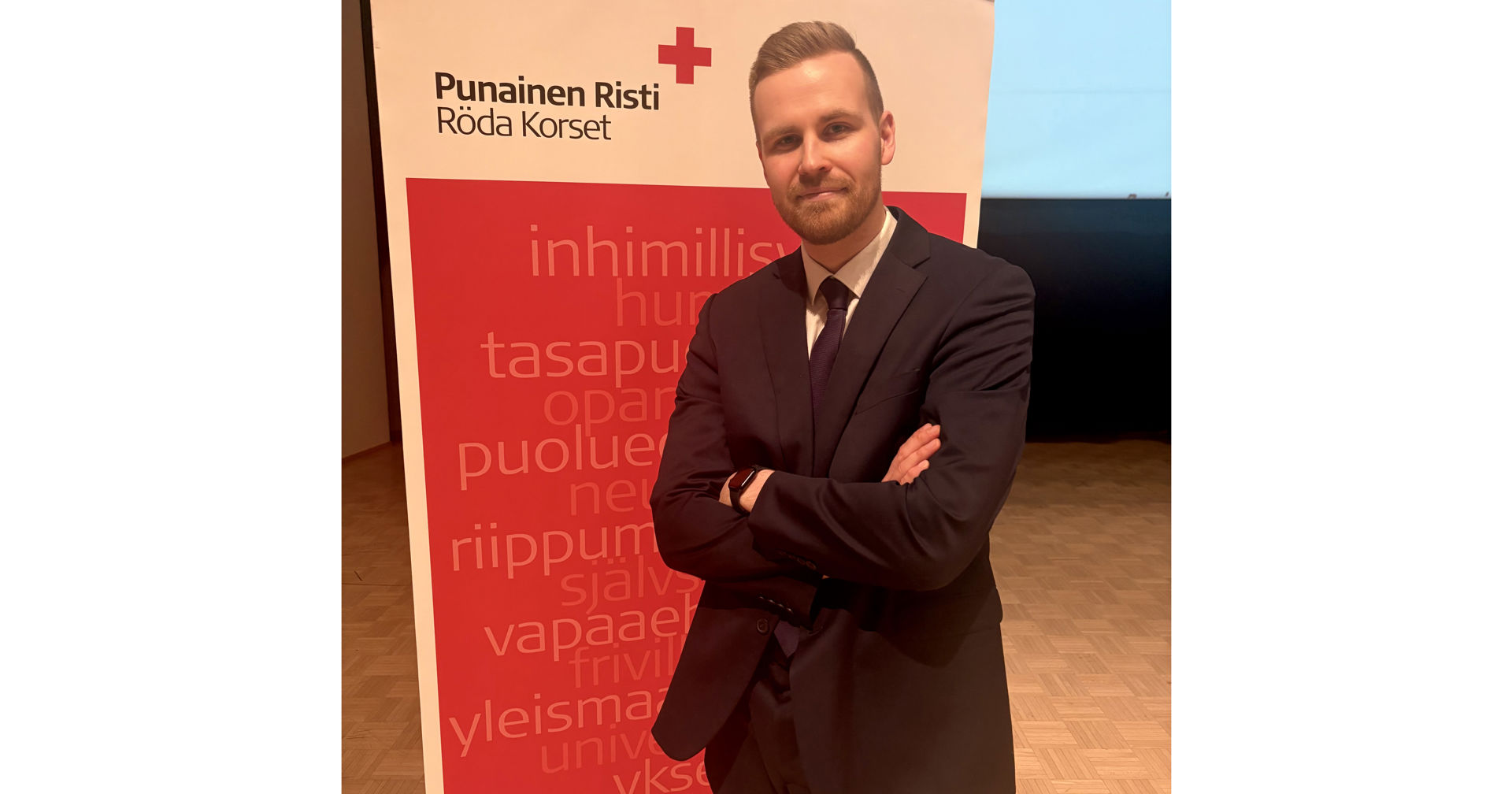 Mikko Sipilä står med armarna i kors framför Röda Korsets banderoll, klädd i mörk kostym och slips.