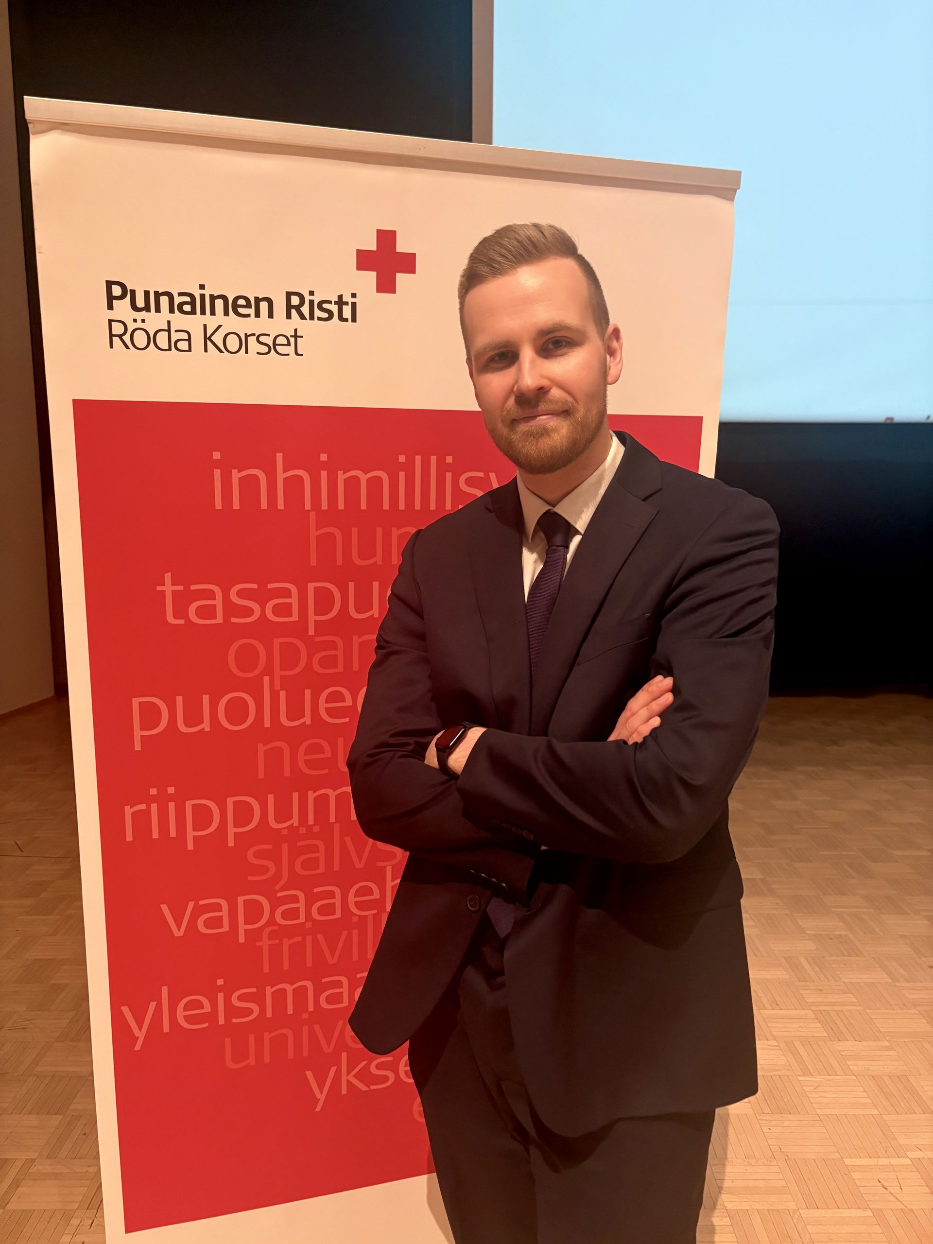 Mikko Sipilä står med armarna i kors framför Röda Korsets banderoll, klädd i mörk kostym och slips.