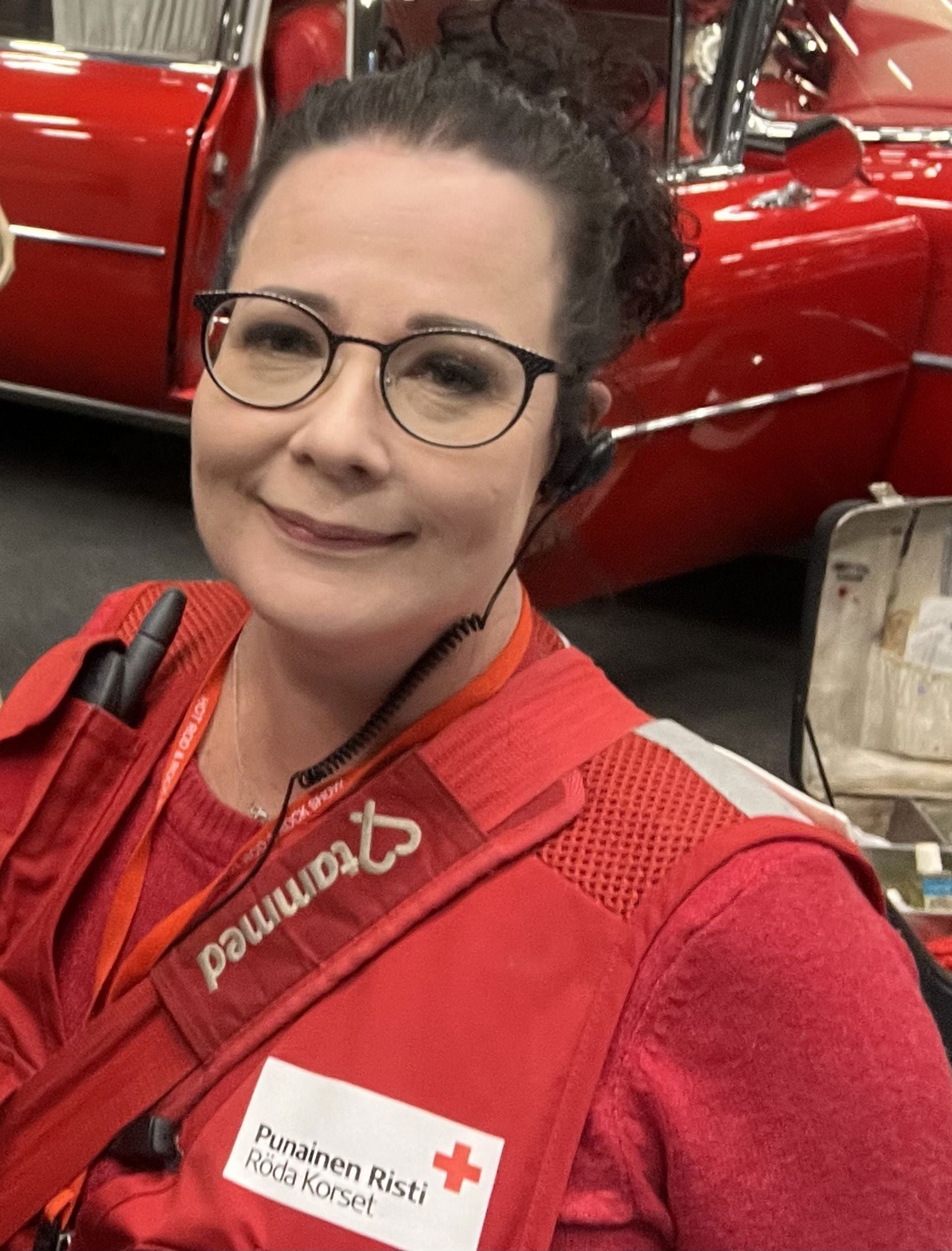 Niina Hokkanen tittar mot kameran, iklädd Röda Korsets väst och glasögon. I bakgrunden finns ett rött fordon.