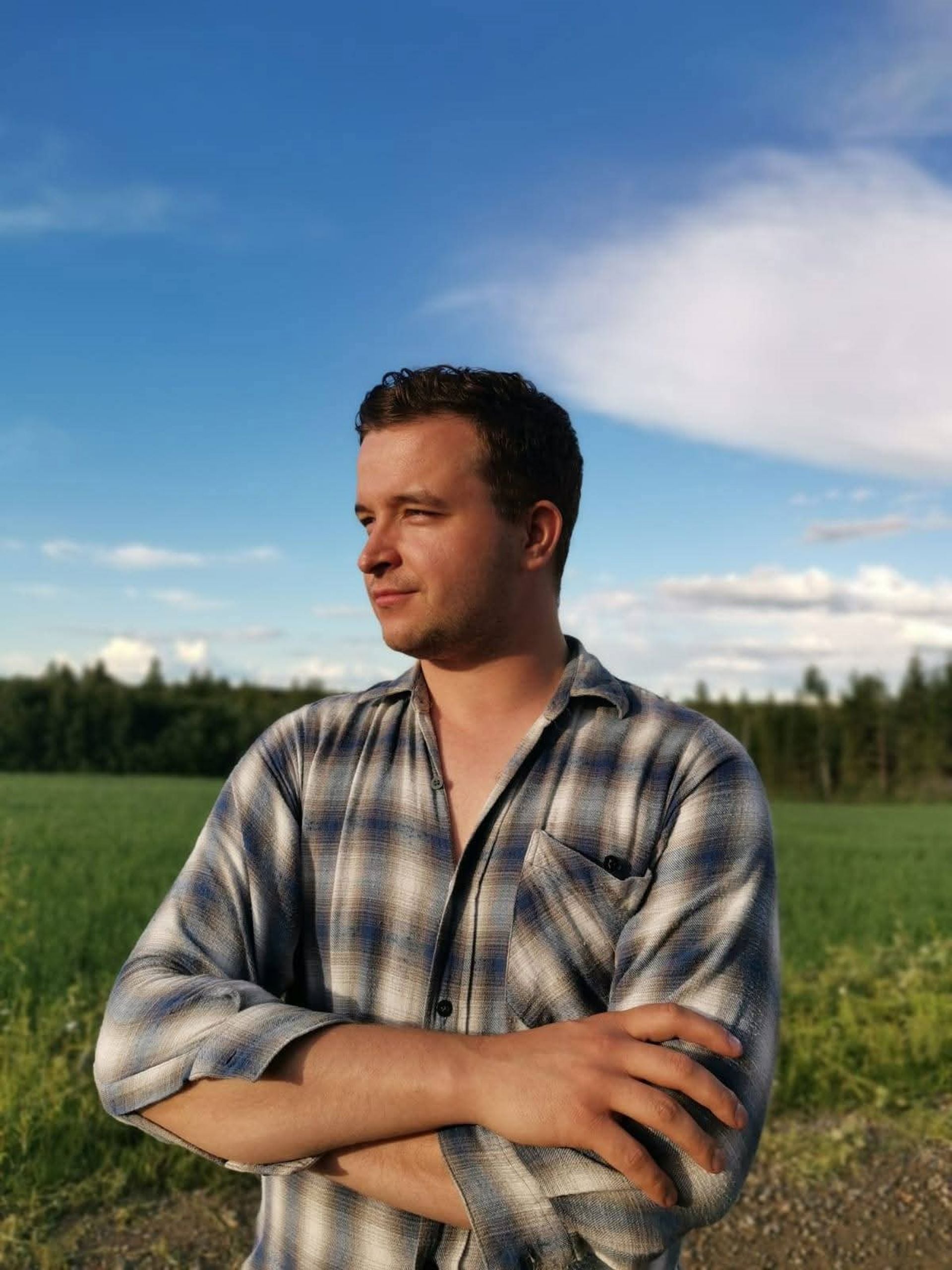 Osmo Tolonen står med armarna i kors vid kanten av en åker, iklädd en rutig skjorta. I bakgrunden syns ett grönt fält, ett skogsbryn och en blå himmel.