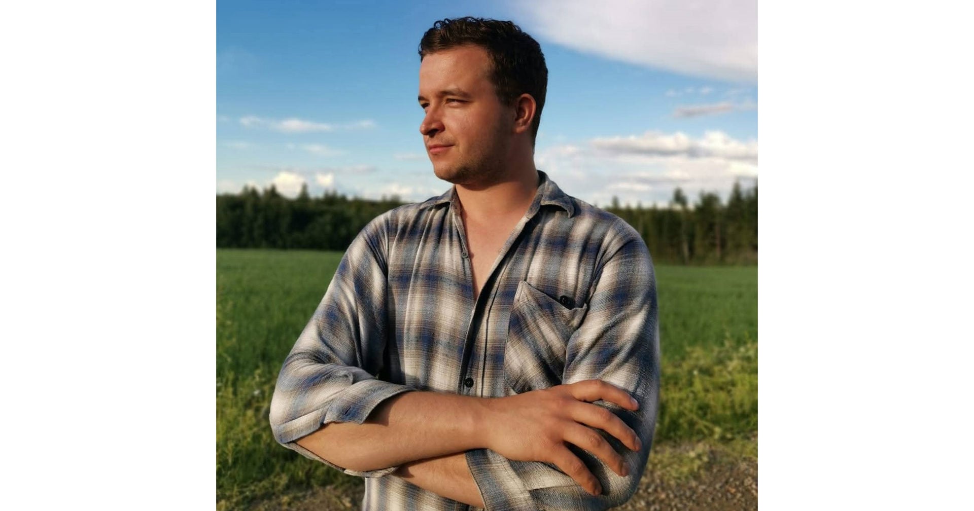 Osmo Tolonen står med armarna i kors vid kanten av en åker, iklädd en rutig skjorta. I bakgrunden syns ett grönt fält, ett skogsbryn och en blå himmel.