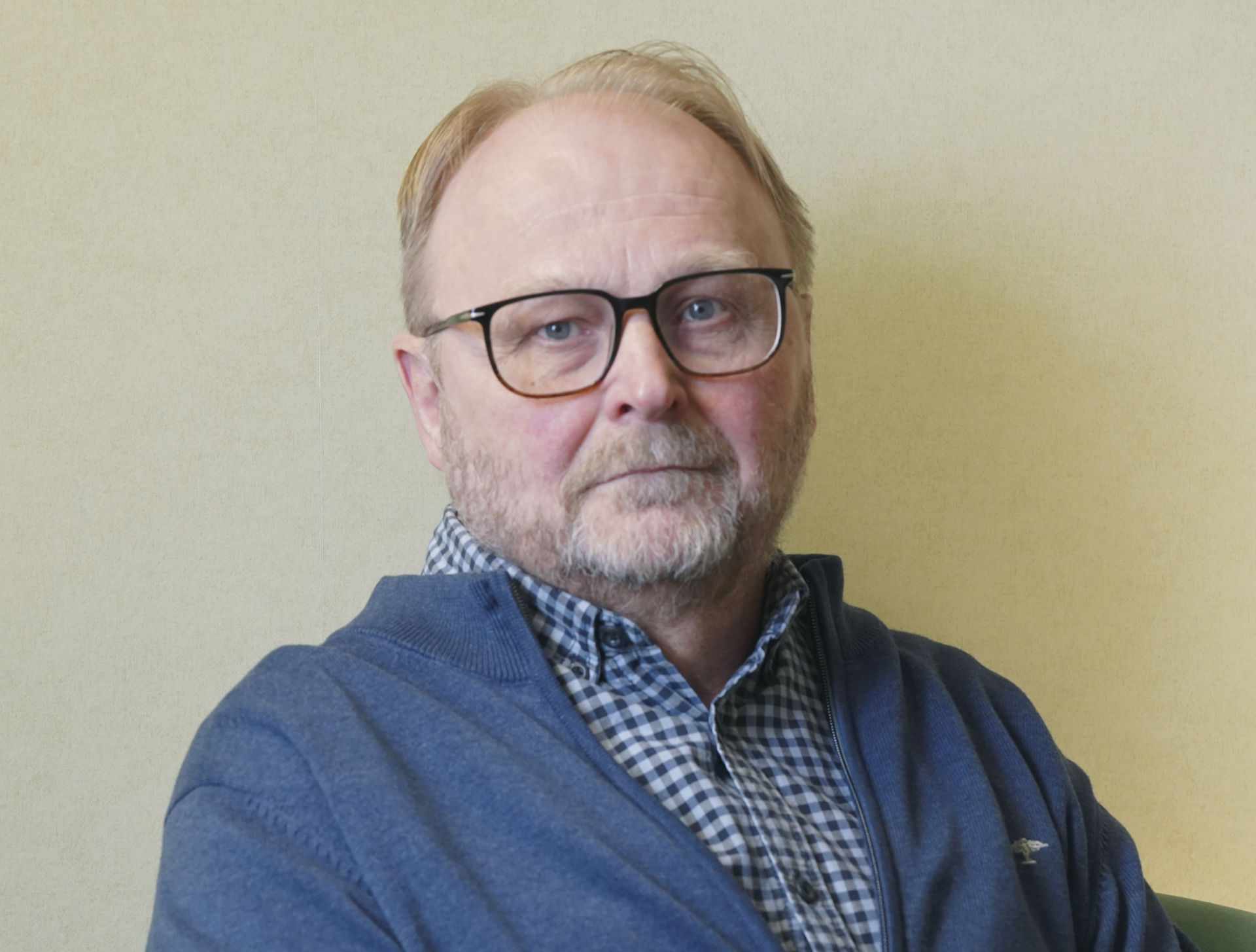 Veli‑Matti Ahtiainen tittar mot kameran, klädd i en rutig skjorta, blå kofta och glasögon.