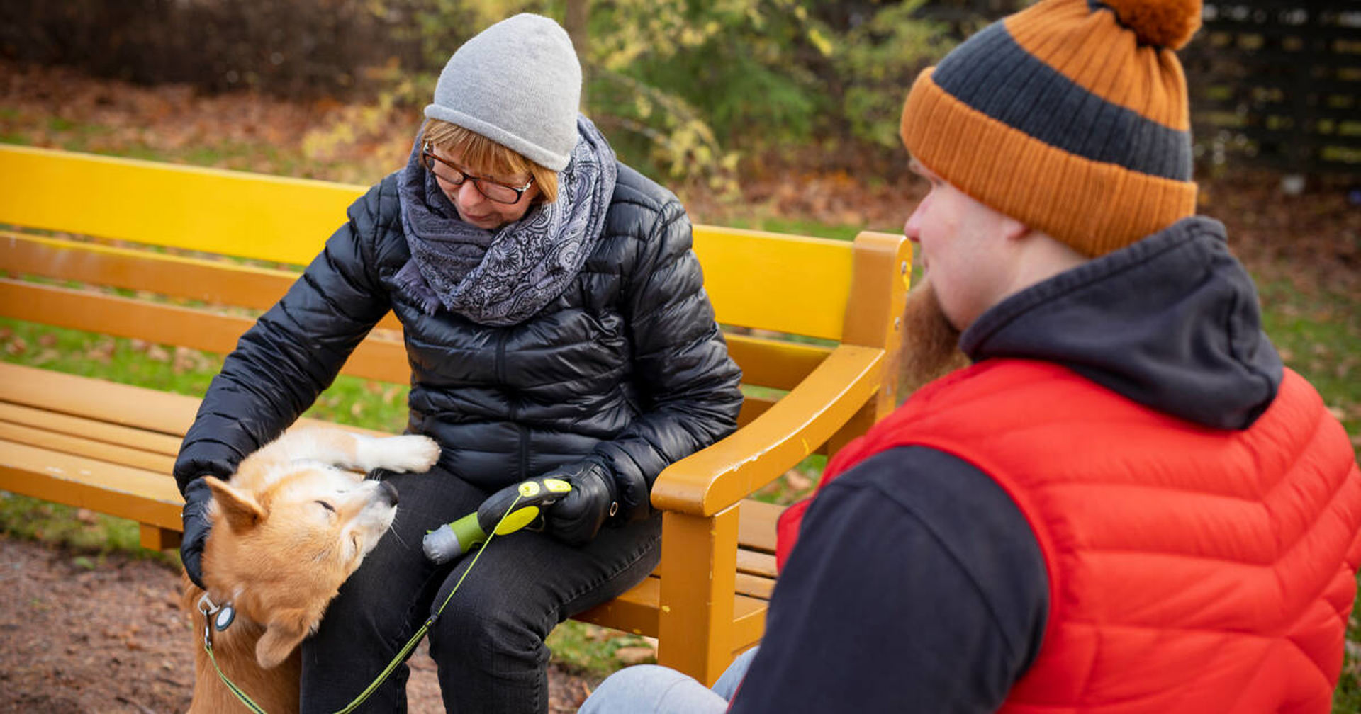 Två personer sitter utomhus. Den ena sitter på en bänk och den andra i en rullstol. De har också en hund.